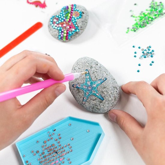 Kinderhanden bevestigen een blauwe zeester van strass steentjes op een grijze steen met een pen.