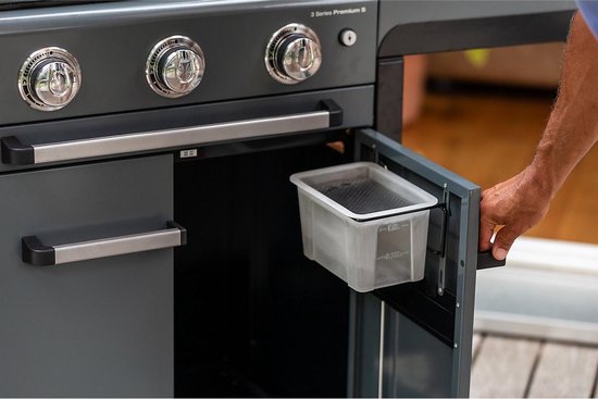 Hand opent de deur van een grijze barbecue, met een transparante opvangbak zichtbaar.