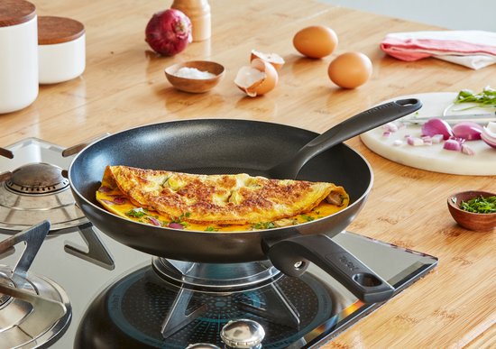 Zwarte koekenpan met een omelet op een gasfornuis. Rode ui en ingrediënten voor ontbijt liggen op het aanrecht.