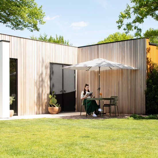 Een vrouw met bruin haar, gekleed in een witte top en lange groene rok, zit aan een ronde tafel onder een grote beige parasol. Ze leest een boek.