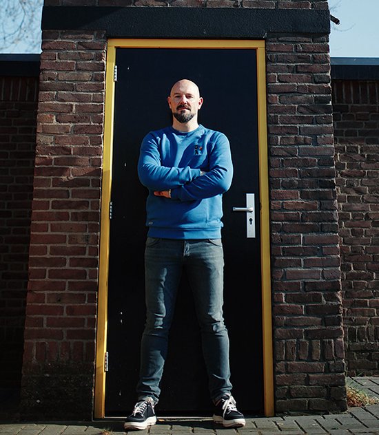 Man met gekruiste armen, staand voor een zwarte deur, draagt een blauwe sweater.