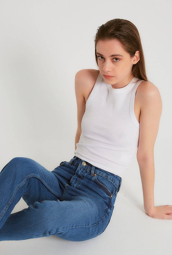 Model zittend in een witte, geribbelde tanktop en donkerblauwe spijkerbroek.