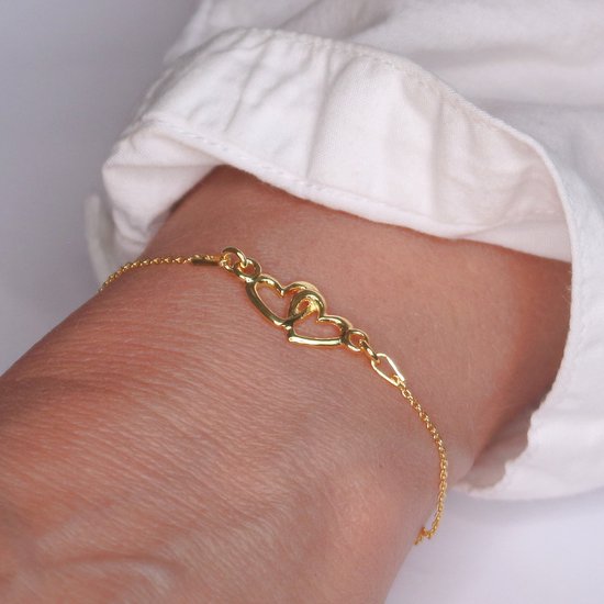 Gouden armband met twee in elkaar verstrengelde hartjes, gedragen op een pols.