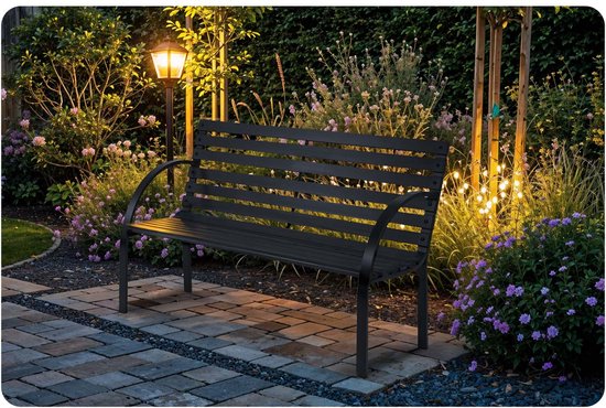 Zwarte houten tuinbank in een tuin bij nacht, verlicht door een lantaarnpaal en decoratieve lampjes.