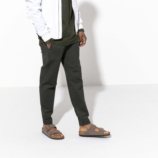 Man met bruine Birkenstock Arizona slippers, grijze broek en wit jack.
