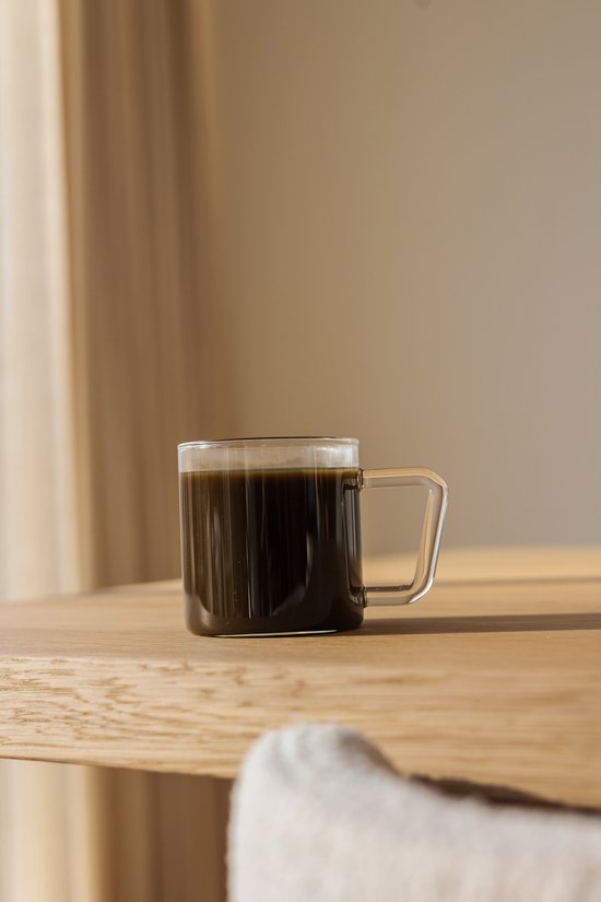 Doorzichtige mok met een oor, gevuld met donkere koffie, staand op een houten tafel.