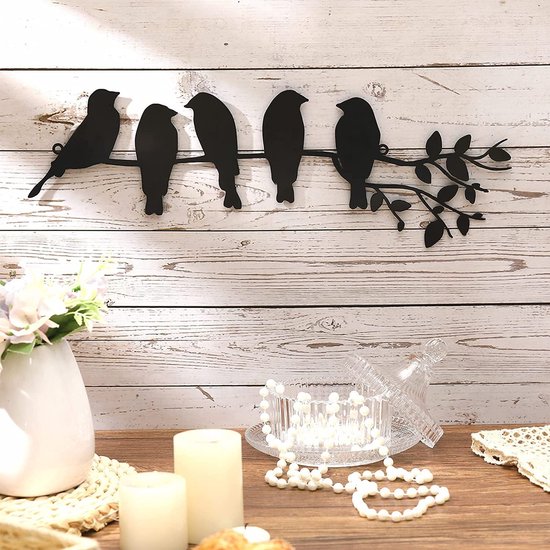 Zwarte metalen wanddecoratie met vijf vogels op een tak, boven een vaas met bloemen.