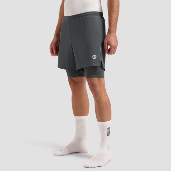 Man met grijze 2-in-1 hardloopshorts, witte sokken en witte sportschoenen. Staande pose.