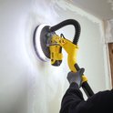 Gele schuurmachine met schijnwerper schuurt een muur. Een persoon met grijze handschoenen en een bril bedient de machine.