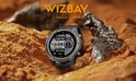Zwarte smartwatch met grijze lunette, midden in een woestijnlandschap. Het scherm toont datum en tijd.