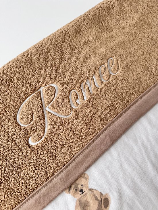 Détail d'un bord en tissu éponge marron avec le nom 'Ranee' brodé en lettres blanches, sur une couverture blanche à imprimé ours en peluche.