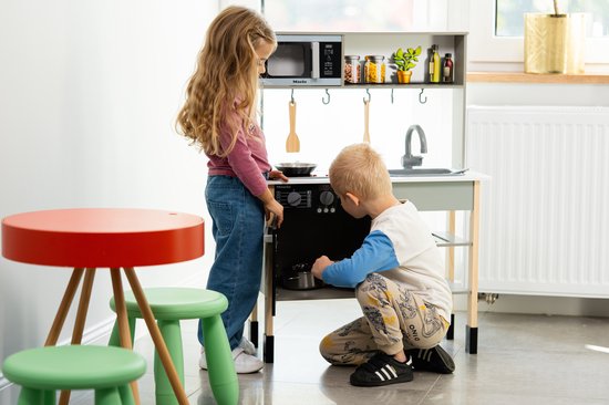 Jongen en meisje spelen bij een witte houten speelkeuken, met een rode tafel en groene krukjes.