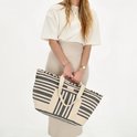 Model met beige rok en wit T-shirt draagt grote canvas strandtas met beige en zwart gestreepte print.