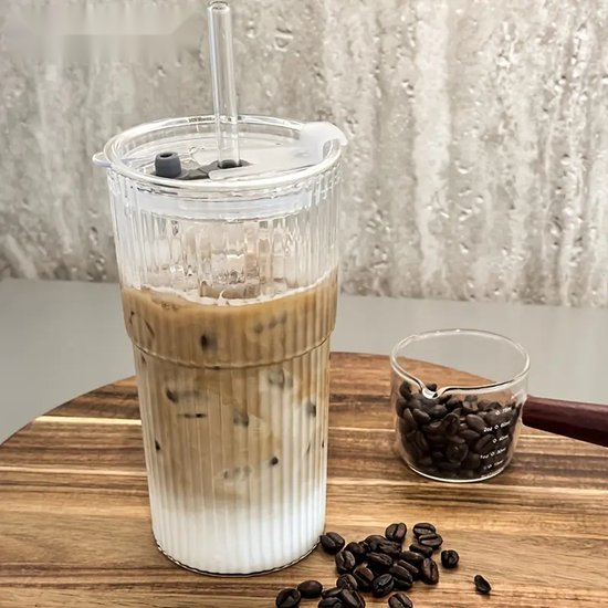 Verre en côtes en verre, rempli de café glacé et d'une paille. À côté du verre, une petite tasse à mesurer en verre avec des grains de café.