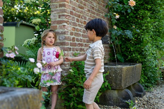 Meisje met blonde vlechtjes draagt een lila jurkje met bloemenprint, een roze bloem in haar hand.