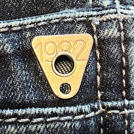 Gouden driehoekige knoop met "1982" op een blauwe spijkerbroek.
