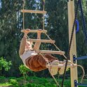 Een meisje met blonde haren hangt aan een touwladder met houten sporten, buiten in het groen.