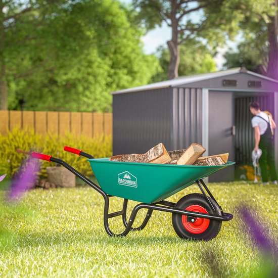 Groene kruiwagen gevuld met houtblokken in een tuin, met een schuur op de achtergrond.