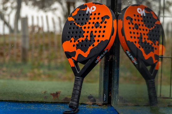 Padelracket met oranje en zwart camouflagepatroon, rustend tegen een glazen wand met reflectie.
