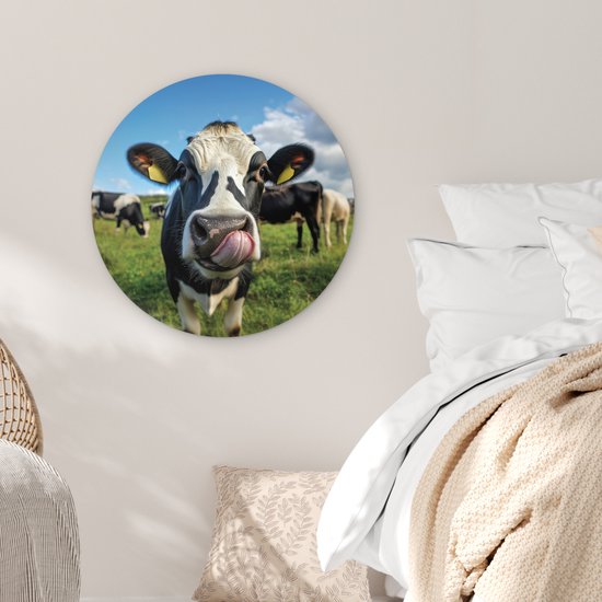 Ronde muurdecoratie met een koe die met haar tong likt op een groen veld onder een blauwe lucht.