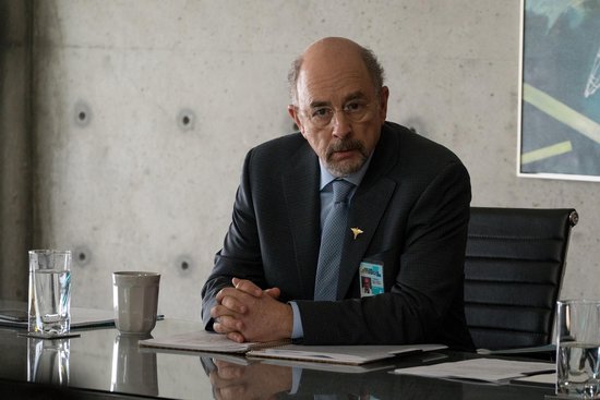 Man met bril en grijs pak met stropdas zit aan een tafel met opgevouwen handen.