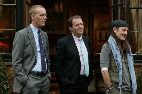 Drie mannen buiten voor een gebouw. Twee in pak, één met bandana en vest.