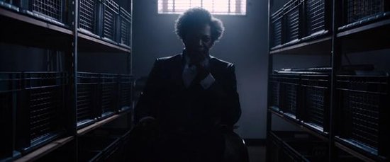 Samuel L. Jackson in een pak, met krullend haar, zit in een donkere kamer tussen metalen rekken, verlicht door een raam.