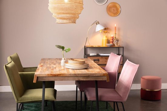 Eettafel met houten blad en zwarte poten, omringd door groene en roze fluwelen stoelen. Een ronde, tweekleurige poef staat rechts.