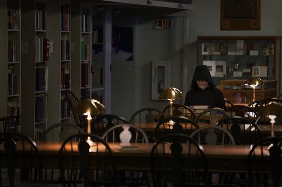 Een figuur in een zwarte hoodie zit aan een tafel in een bibliotheek met lamplicht.