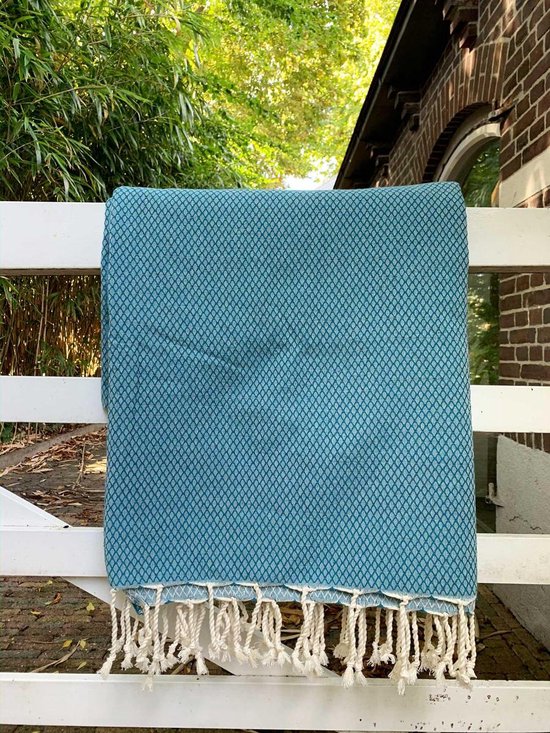 Petrolblauwe plaid met ruitpatroon en beige franjes, hangend over een witte schutting.