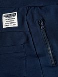 Detail van een donkerblauwe joggingbroek met een label met tekst, een zak en een ritssluiting.