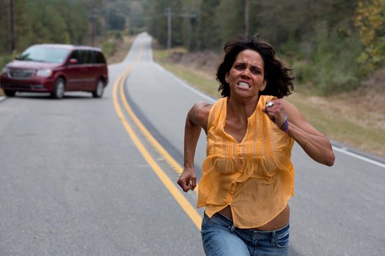 Halle Berry rent met een grimmige uitdrukking op een weg, met een auto op de achtergrond.