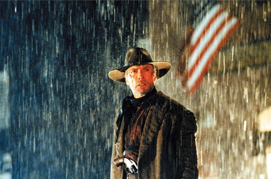 Clint Eastwood met een cowboyhoed en regenjas, staande in de regen, met een Amerikaanse vlag op de achtergrond.