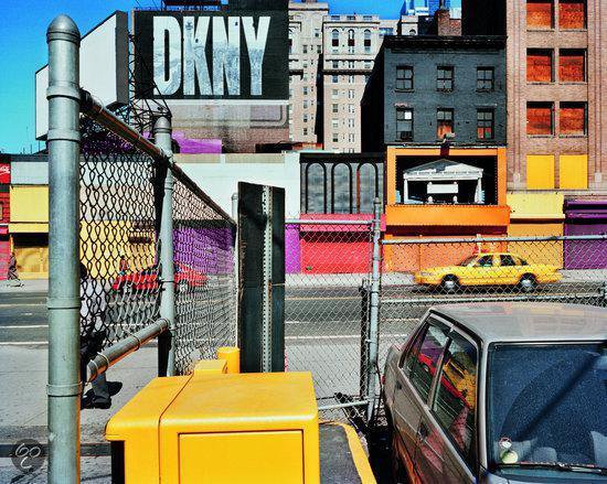 Stadsgezicht met DKNY reclame, felgekleurde gebouwen, hekwerk, gele auto en zilverkleurige auto.