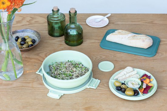 Boîte à salade vert pâle sur une table avec du pain, des légumes et des olives.
