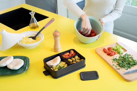 Personen bereiden lunch in een keuken met een donkergrijze bento lunchbox met pitabroodje, olijven en tomaten.