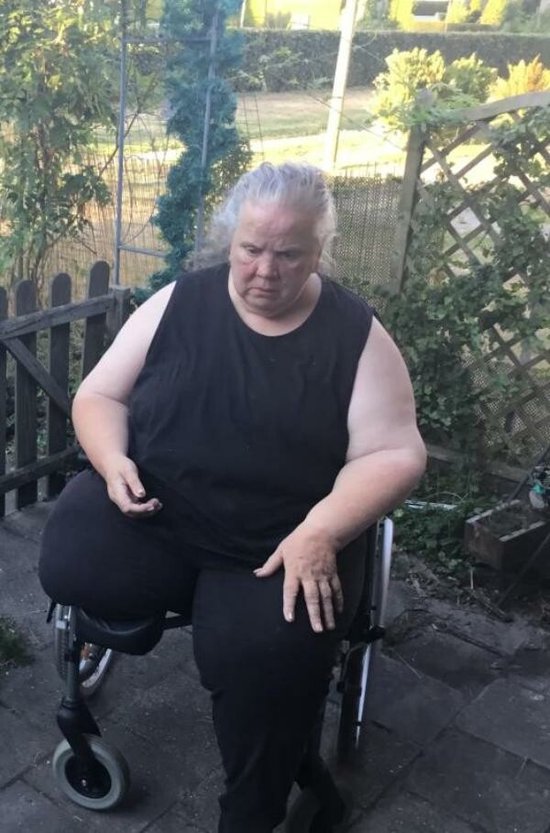 Volle vrouw met grijzend haar, gekleed in een zwart T-shirt en zwarte broek, zit in een rolstoel in een tuin.