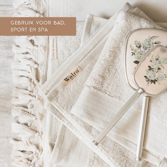 Crèmekleurige handdoeken met franjes en labels 'Walra' en 'LOVE THE SOFTNESS'. Een sierlijke spiegel ligt erop.