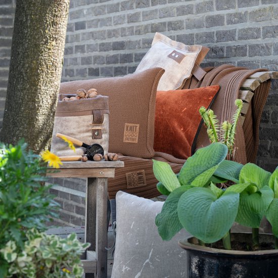 Een bruine gebreide plaid en kussens op een houten bank buiten, met een theepot en planten.