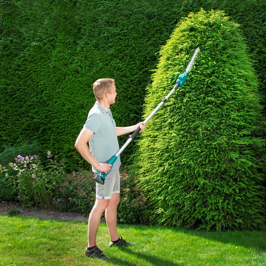 Man snoeit een hoge groene heg met een lange, groene telescoop heggenschaar op een zonnige dag.