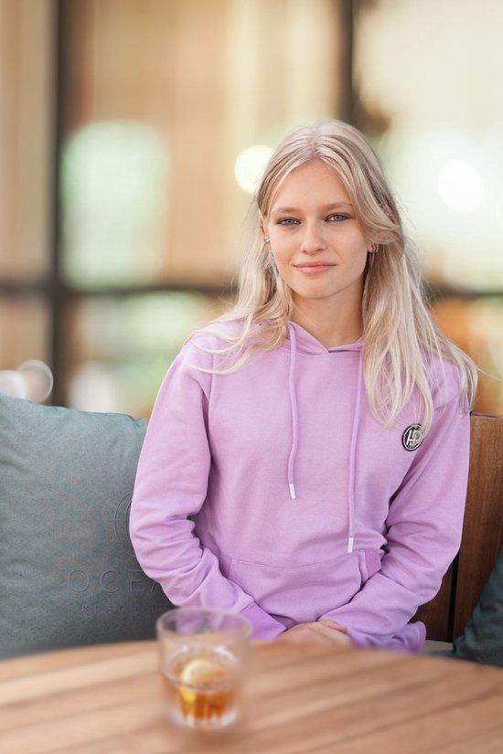 Vrouw zit op een bankje in een zachtpaars joggingpak met capuchon en een glas met drankje op tafel.