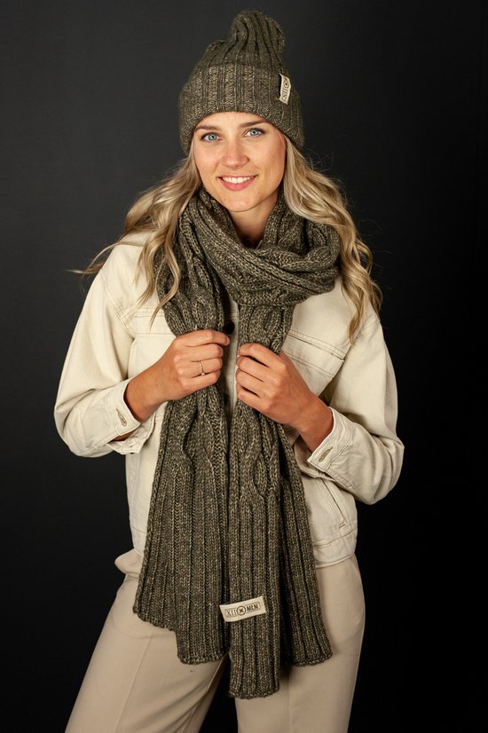 Model met een gebreide donkergroene muts en sjaal met kabelpatroon. Ze draagt een beige jas.