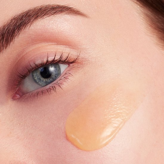 Een druppel gelige crème wordt op een lichte huid aangebracht, naast een oog.