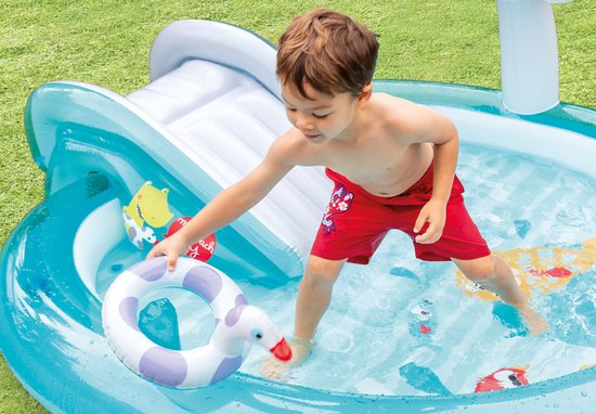 Jongen met rode zwembroek pakt badeendje in opblaasbaar zwembad met glijbaan.