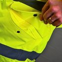 Buitenkant van een felgele werkjas met twee voorzakken met drukknoopsluiting. Een hand opent de rechterzak.