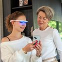 Vrouw met witte bril met blauwe lampjes kijkt op smartphone, naast oudere vrouw.