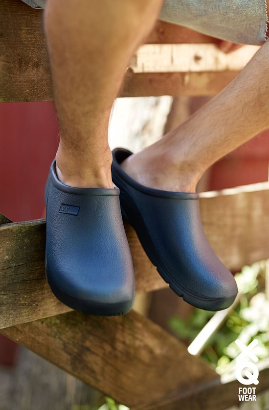 Voeten van een man in donkerblauwe tuinklompen, zittend op een houten hek.