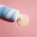 Fles hydraterende haarreiniger met blauwe en witte strepen ligt op zijn kant, met product dat erop druppelt op een roze achtergrond.