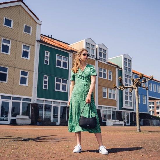 Model in groene jurk met bloemenprint, witte sneakers, zwarte tas en zonnebril voor kleurrijke gebouwen.