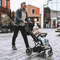 Man met bril en groene jas duwt een zwarte Deryan buggy met een baby erin, buiten.